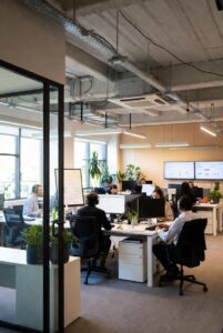 Levende co-working-rum i en innovationshub med teams, der samarbejder ved fælles skriveborde, flere store skærme, der viser datavisualiseringer, synlige lofter og frodige indendørsplanter